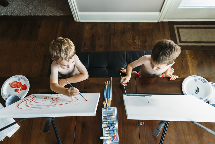 Overhead view of shirtless boys painting on canvas at home