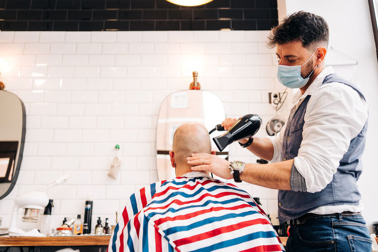 Anonymous male stylist in mask using hairdryer while touching bald head of man in striped cloth in beauty salon