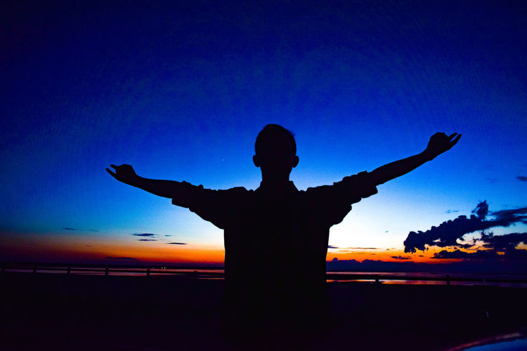 Silhouette man with arms outstretched standing against sky during sunset