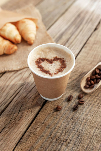 Paper cup of coffee with heart shape, cream, foam, croissants bundle at sackcloth, dried beans