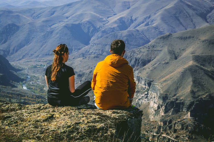 Rear view of couple sitting on cliff against mountains