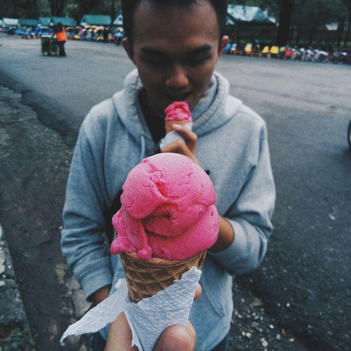 Cropped hand holding strawberry ice cream cone in front of man in city