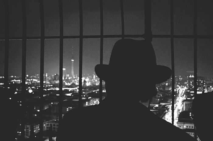 Rear view of man looking at illuminated cityscape from metal bars at night