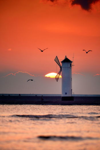 Silhouette birds flying over sea against orange sky and lighthouse in shape of a mill