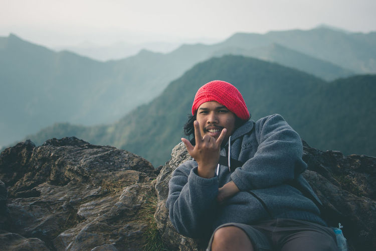 Portrait of man gesturing while leaning on rock against mountains