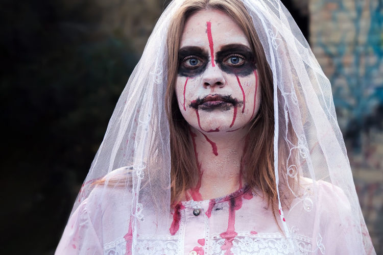 Portrait of zombie woman in wedding dress with veil and stage makeup looking into camera. 