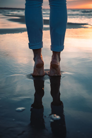 Low section of man standing on sea shore against sky during sunset