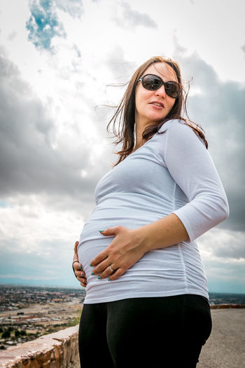 Pregnant woman with hands on stomach standing against cloudy sky