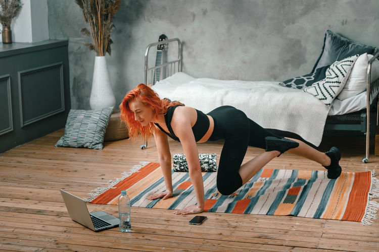 Positive athletic woman with red hair and tight sportswear lunges at home. 