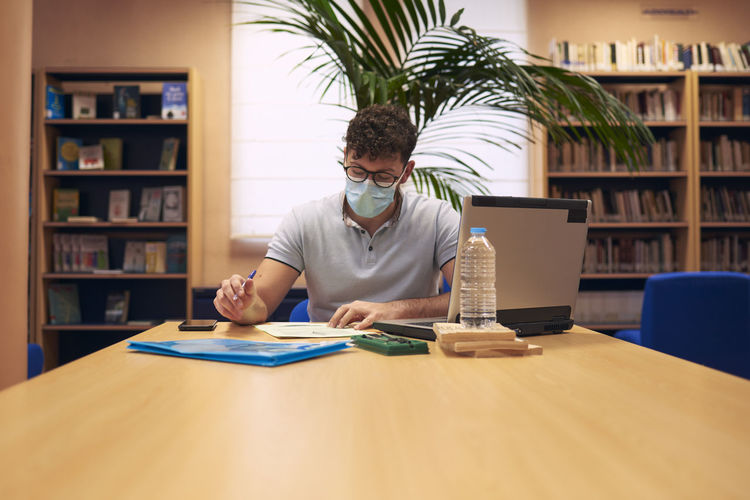 A young boy with a mask sitting and working with his laptop in the library