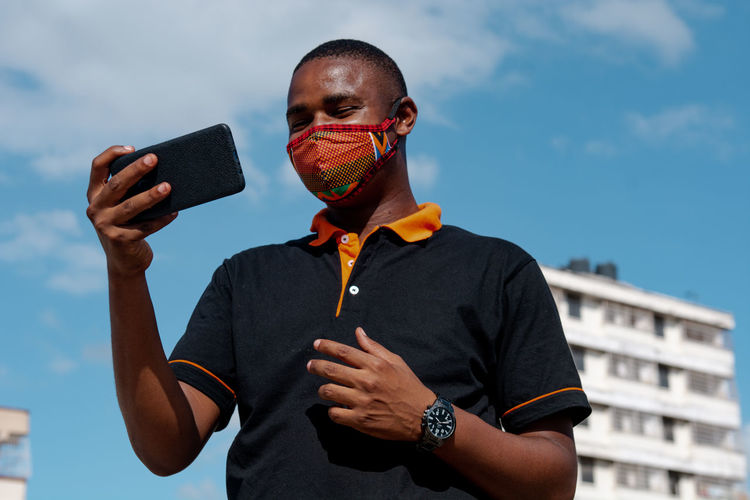 Low angel view of man wearing flu mask holding smart phone against sky
