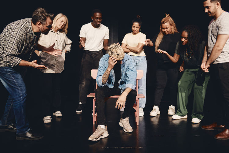 Distraught man scratching head while sitting amidst angry multiracial actors on stage