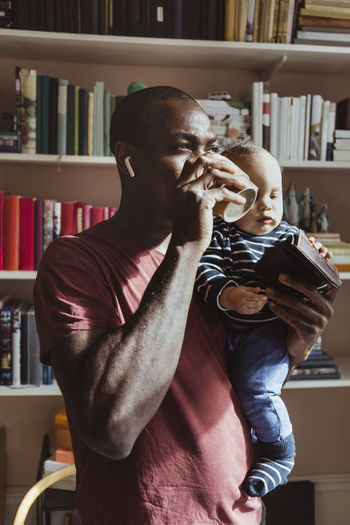 Father drinking coffee while carrying male toddler at home