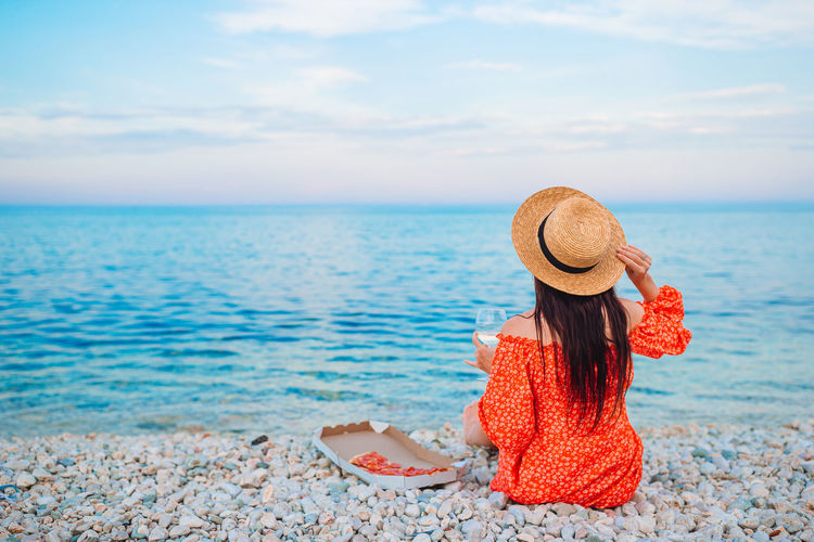 Rear view of woman wearing hat sitting on beach against sky