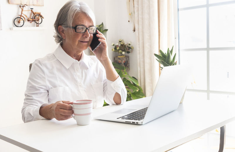 Smiling senior woman holding coffee cup talking on phone while sitting at office
