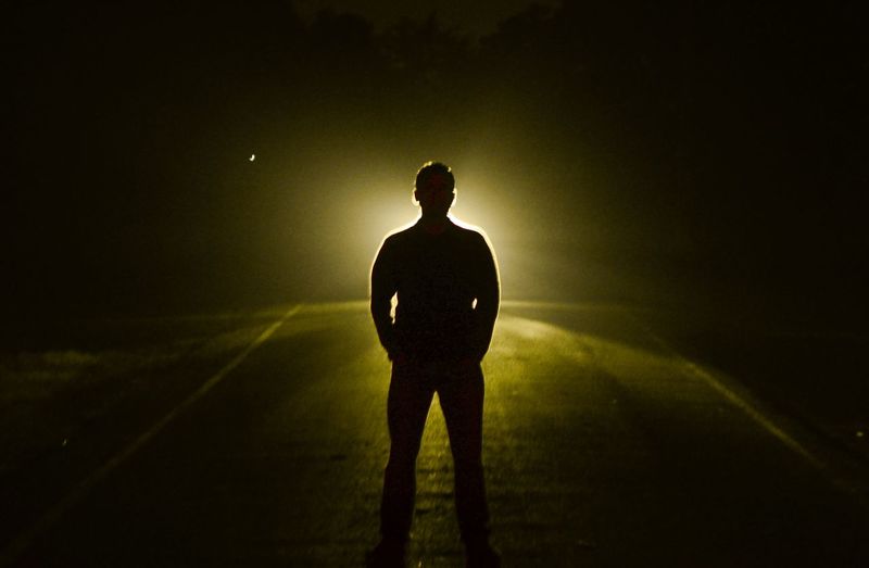 Silhouette man standing on middle of road against vehicle at night