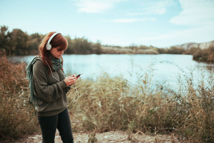 Young woman listening to music with headphones near a lake with her smartphone