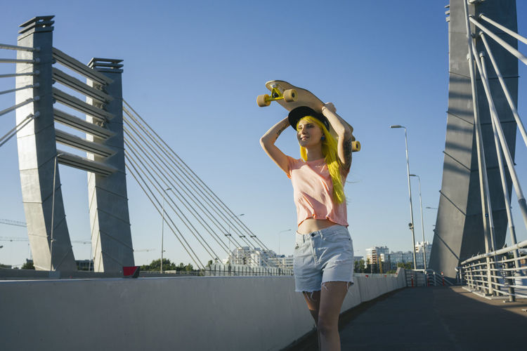 Young woman lifting skateboard on head while standing on bridge during sunny day
