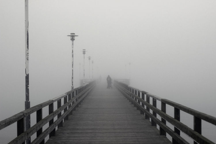 Man walking on footbridge by pier against sky during foggy weather