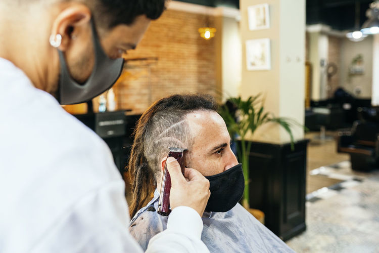 Unrecognizable crop male hairdresser using trimmer and grooming hair of man with dreadlocks in contemporary barbershop