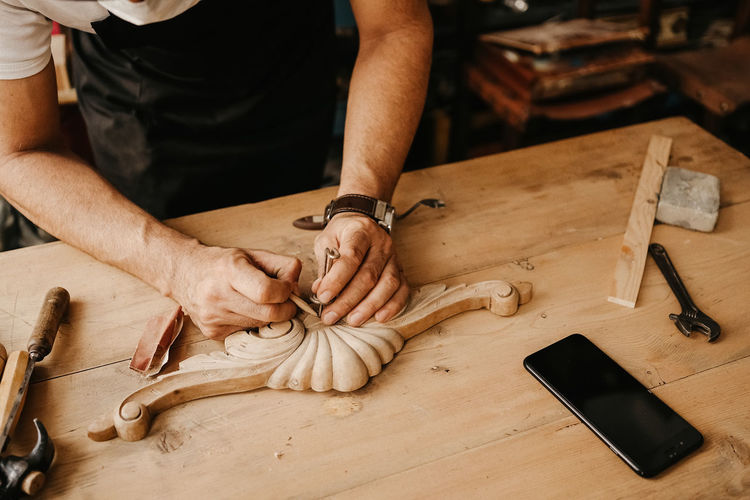 Crop anonymous male master in apron using sharp chisel while creating carved ornate decorative wooden detail at workbench with tools in workshop