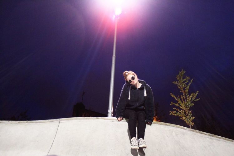Low angle view of woman sitting on retaining wall by illuminated street light against sky at night