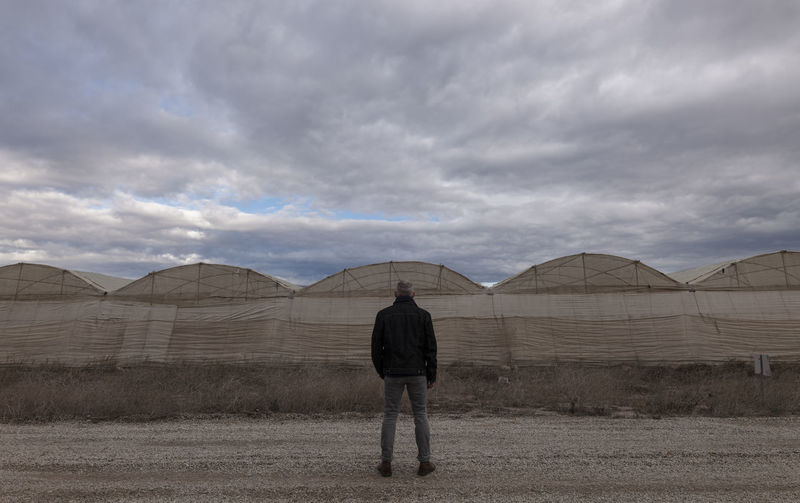 Rear view of adult man in front of plastic greenhouses against cloudy sky in almeria, spain