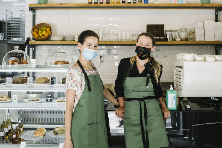 Partners wearing face mask and apron standing by counter at coffee shop