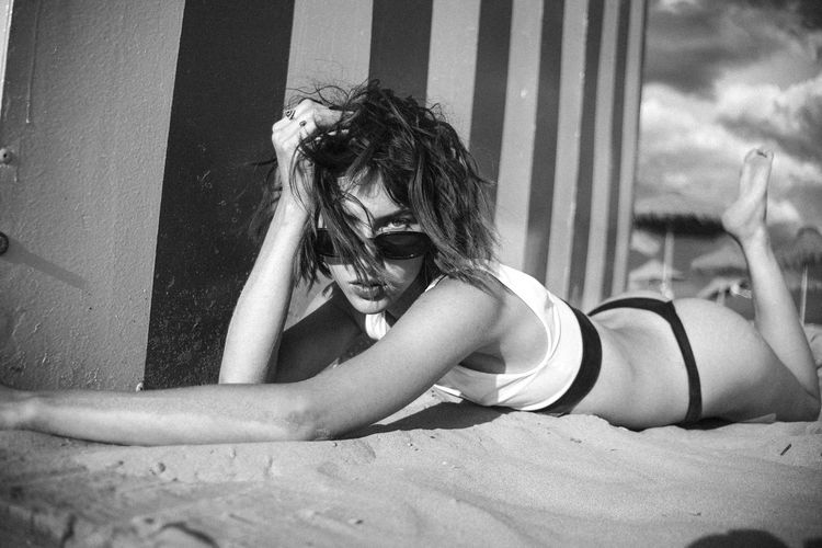 Portrait of young woman wearing bikini and sunglasses lying on sand at beach 