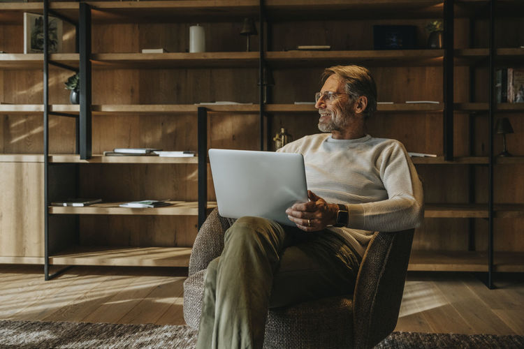 Smiling mature man with laptop sitting on armchair