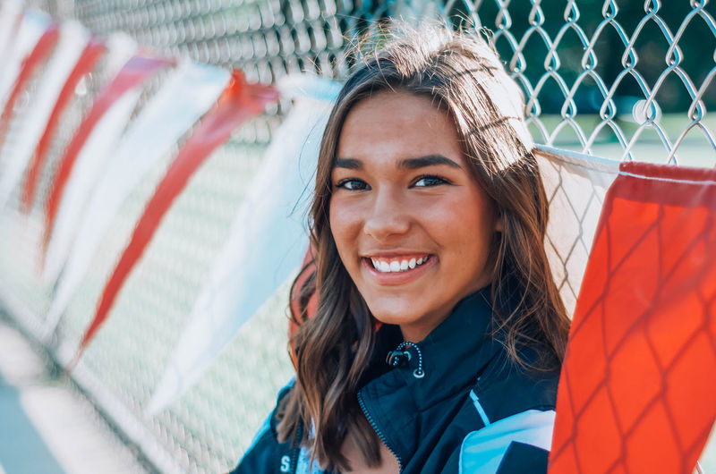 Portrait of smiling young woman sitting by chain-link fence