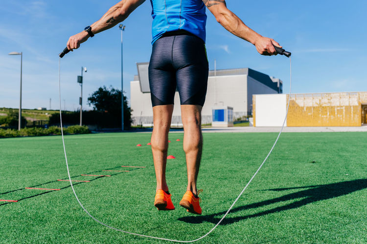 Back view of young male athlete jumping with skipping rope on green lawn during functional workout on street in summer day