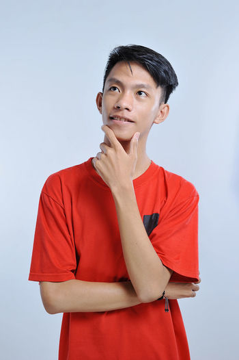 Handsome young asian boy wearing orange t-shirt with hand on chin thinking about question, pensive expression. smiling with thoughtful face. doubt concept