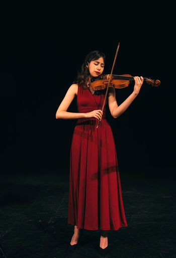 Full body woman in red dress playing violin with closed eyes during concert on stage in dark theater