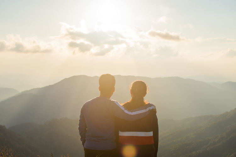 Rear view of couple standing on mountain against sky during sunset