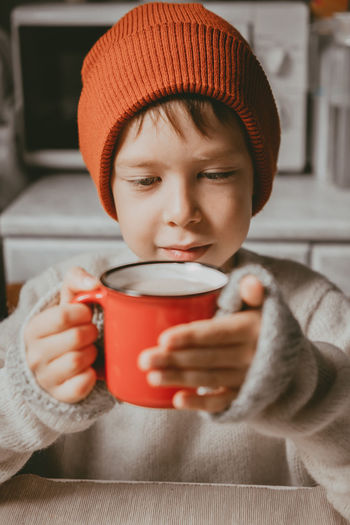 A boy in a sweater and a brown hat drinks cocoa from a red cup. a cozy photo with a mug in hand.