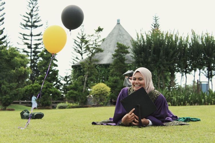 Portrait of happy woman wearing graduation gown relaxing on grassy field 