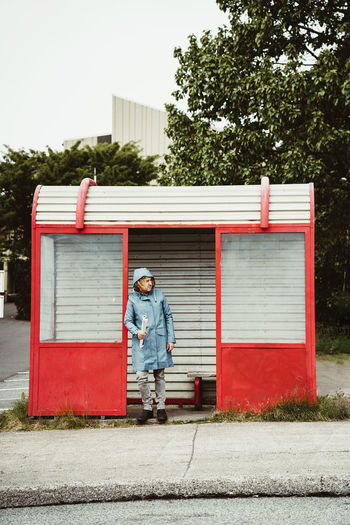 Full body of pensive person in raincoat standing and leaning on wall of bus stop near roadway in town in daylight person