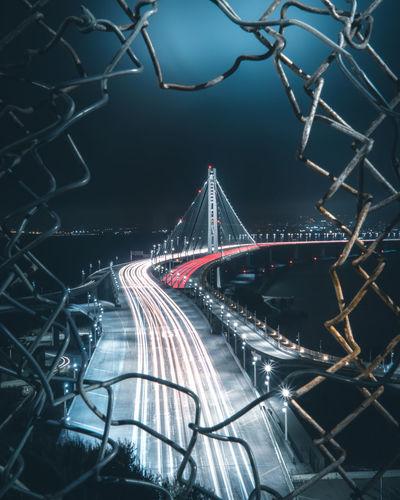 High angle view of light trails on bridge seen through hole in fence