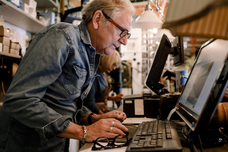 Side view of confident salesman in denim shirt using computer at hardware store checkout