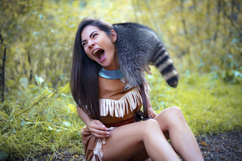 Young woman in traditional clothing playing with raccoon in forest