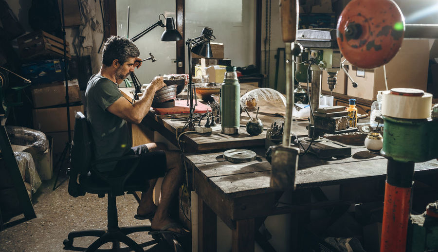 Side view of middle aged craftsman in casual clothes sitting at workbench and creating accessory from silver during work in professional workshop