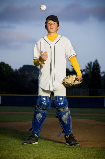 Portrait of a boy standing in white baseball uniform and catchers shin guards tossing a baseball in the air