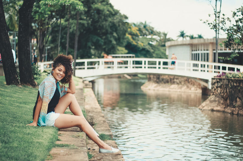 Portrait of young woman sitting by canal against trees and bridge