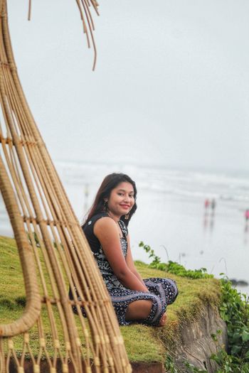 Portrait of smiling young woman sitting on shore by sea against sky