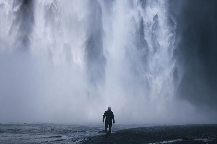 Back view of anonymous male explorer in casual clothes and hat walking near wild river with black sandy shore towards powerful skogafoss waterfall during trip in iceland