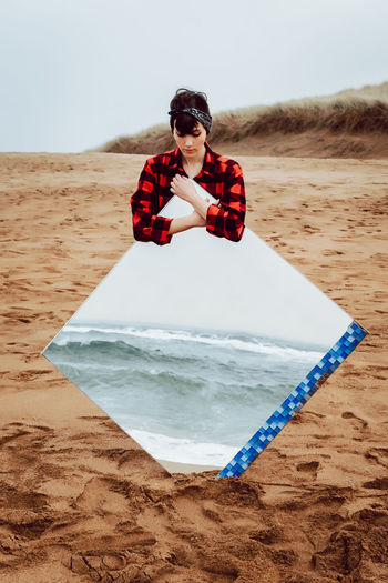Pensive unhappy lonely young female in casual outfit standing on empty sandy beach in gloomy day and holding large mirror with reflection of stormy sea