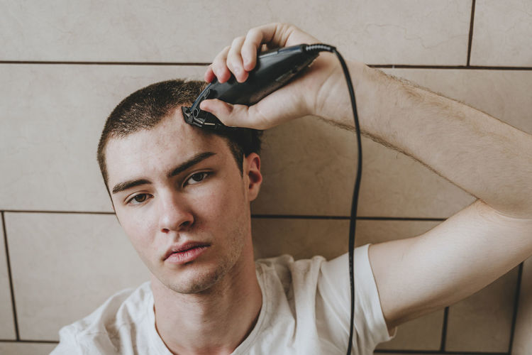 Lonely young man shaving hair with electric razor in bathroom