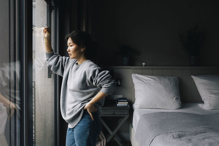 Thoughtful lonely woman looking out through window while standing with hand on hip in bedroom at home