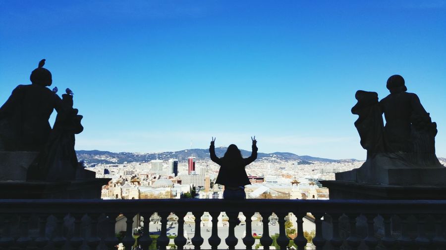 Rear view of woman showing peace sign by cityscape against clear blue sky